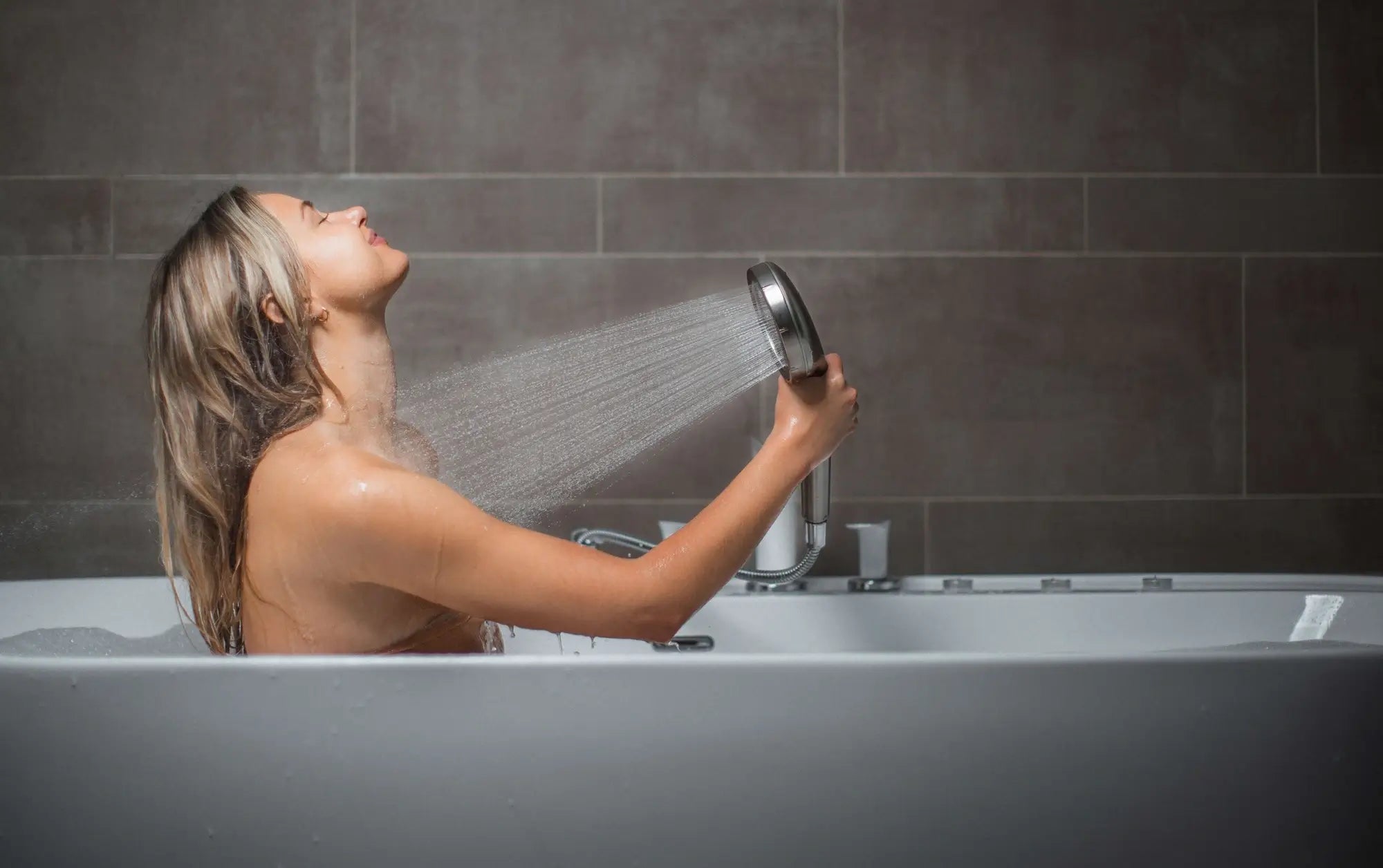 Side-view of blonde female in bathtub showering with blu Ionic Shower Filter Handheld